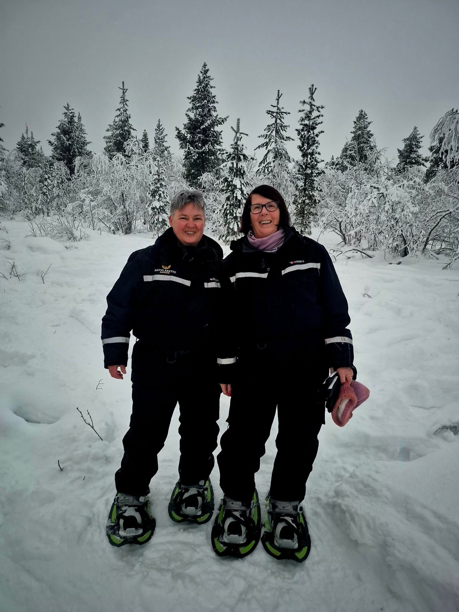 Two women in snowshoes stand in a snowy forest clearing after a winter walk, with frosted pines rising behind them under a grey sky.